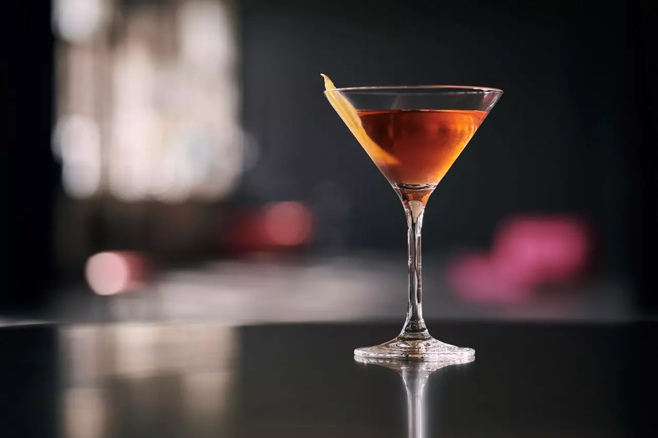 A classic cocktail in a martini glass with an orange peel garnish sits on a reflective surface, with a blurred dark background.