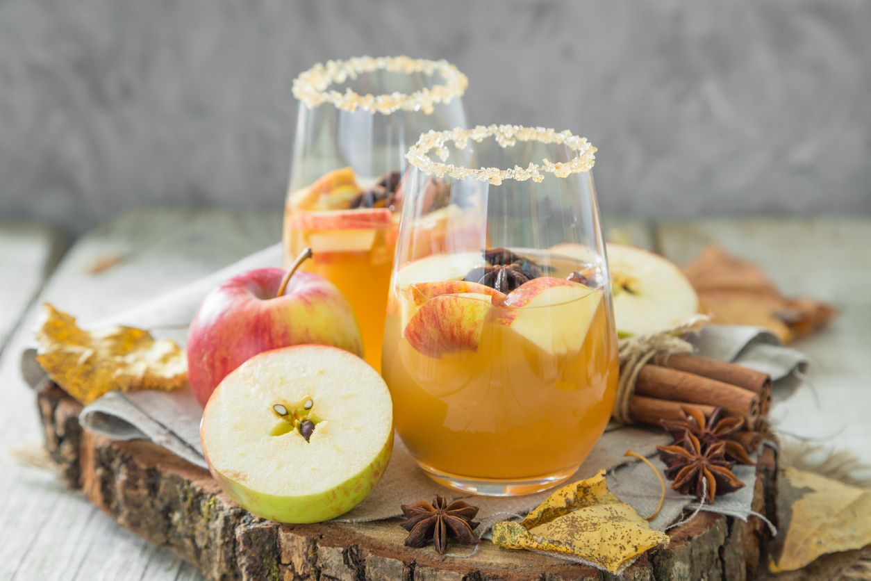 Two glasses of apple cider garnished with apple slices and spices, set on a wooden tray with fresh apples, cinnamon sticks, star anise, and autumn leaves.