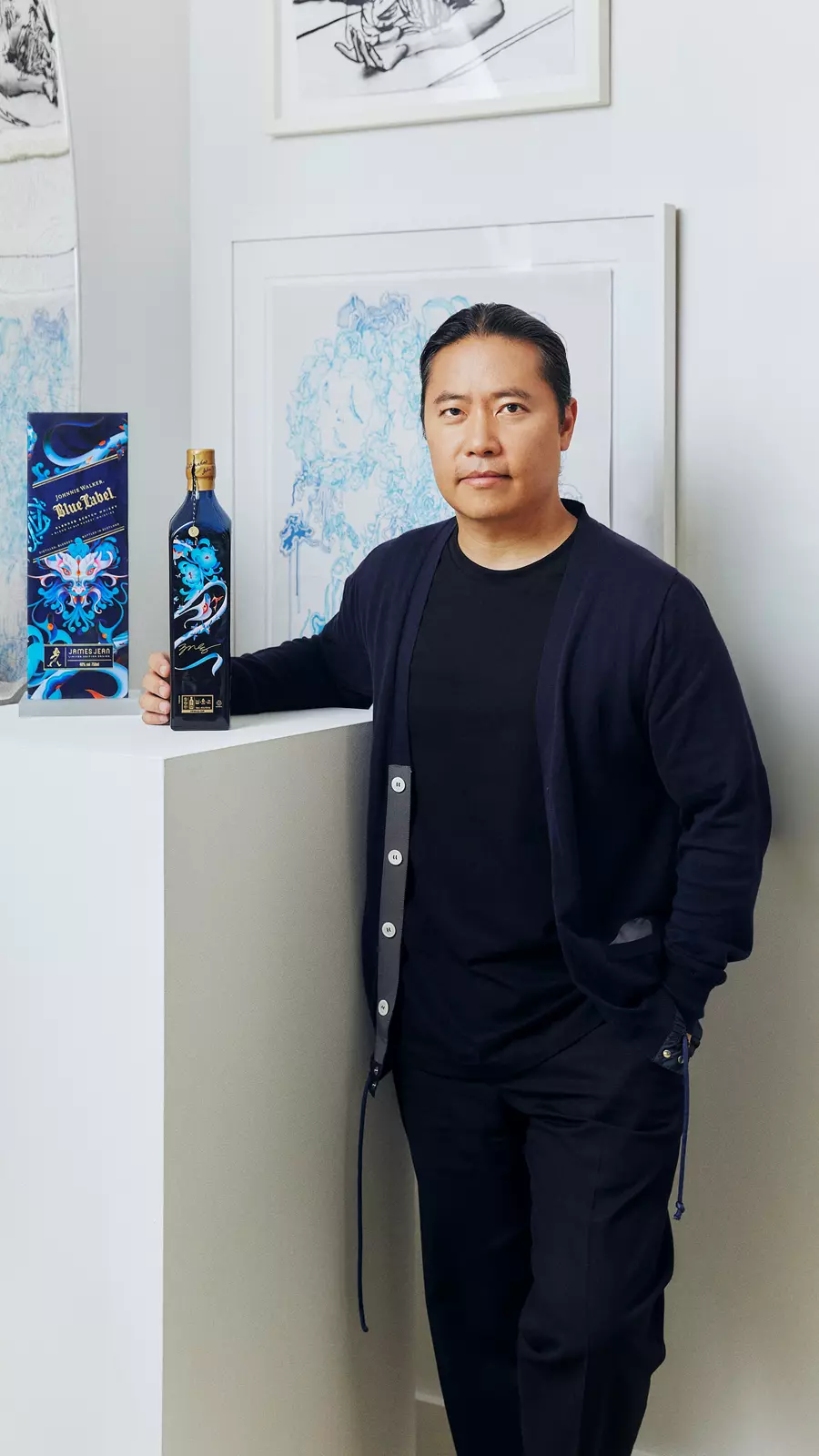 A man in dark clothing stands indoors next to a decorated bottle and its box, with framed artwork on the wall behind him.