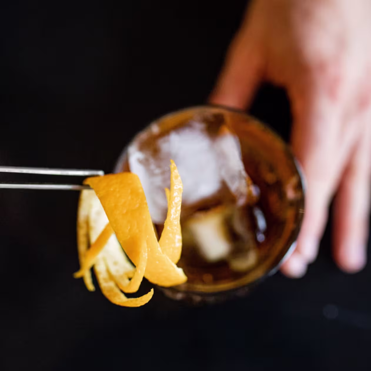 Mano sosteniendo un vaso con hielo mientras se decora con una espiral de piel de cítrico utilizando pinzas de cóctel.