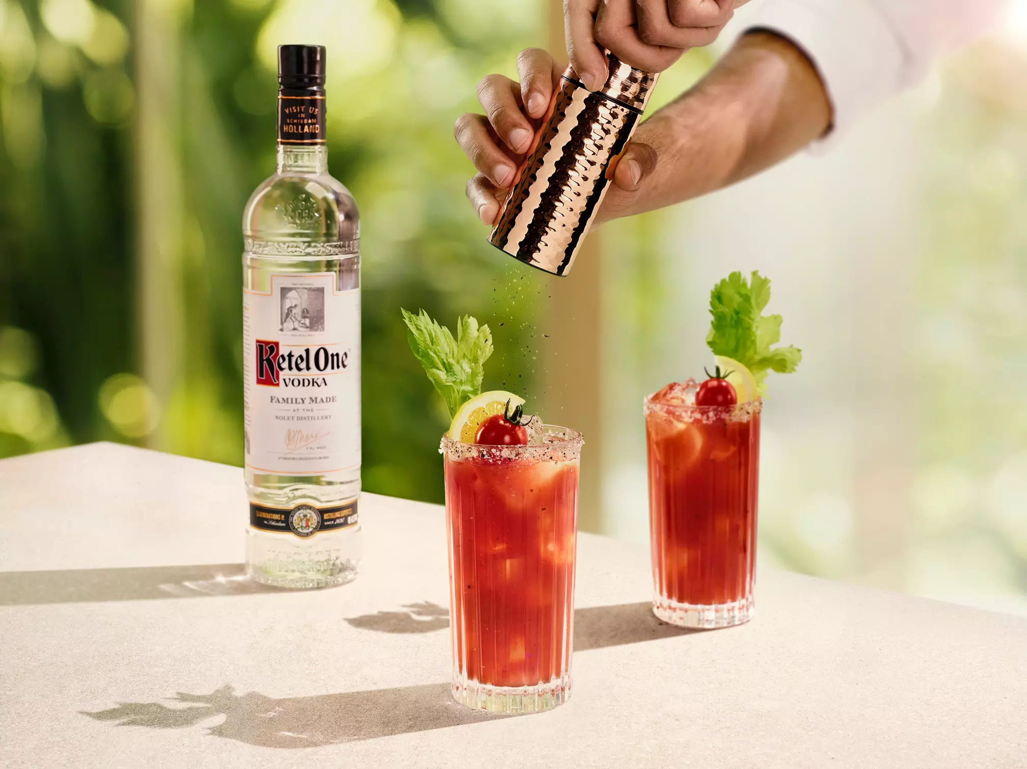 A bottle of Ketel One vodka stands behind two Bloody Mary cocktails garnished with celery, lemon, and tomato; a hand sprinkles seasoning over the drinks.