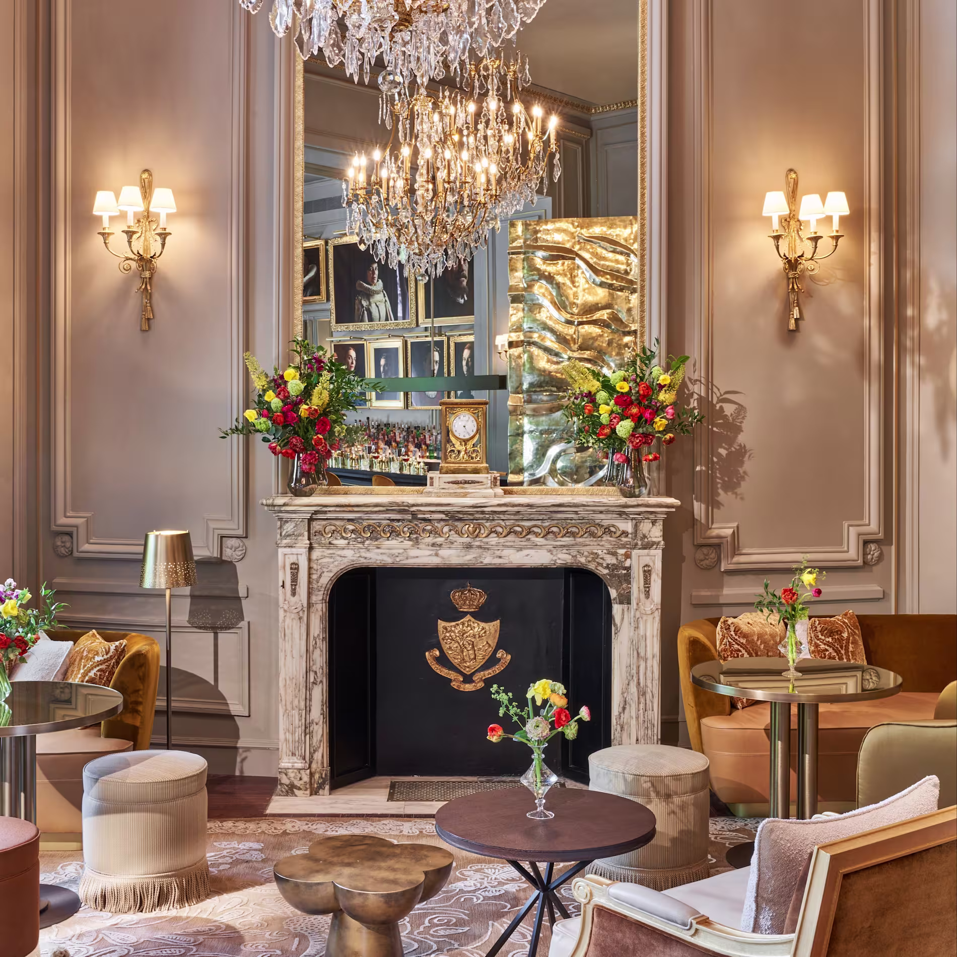 La imagen muestra una habitación del Pictura Elegantemente decorada. El punto focal es una chimenea con un espejo ornamentado encima, flanqueada por luces de pared. La repisa está adornada con vegetación y flores rojas. 