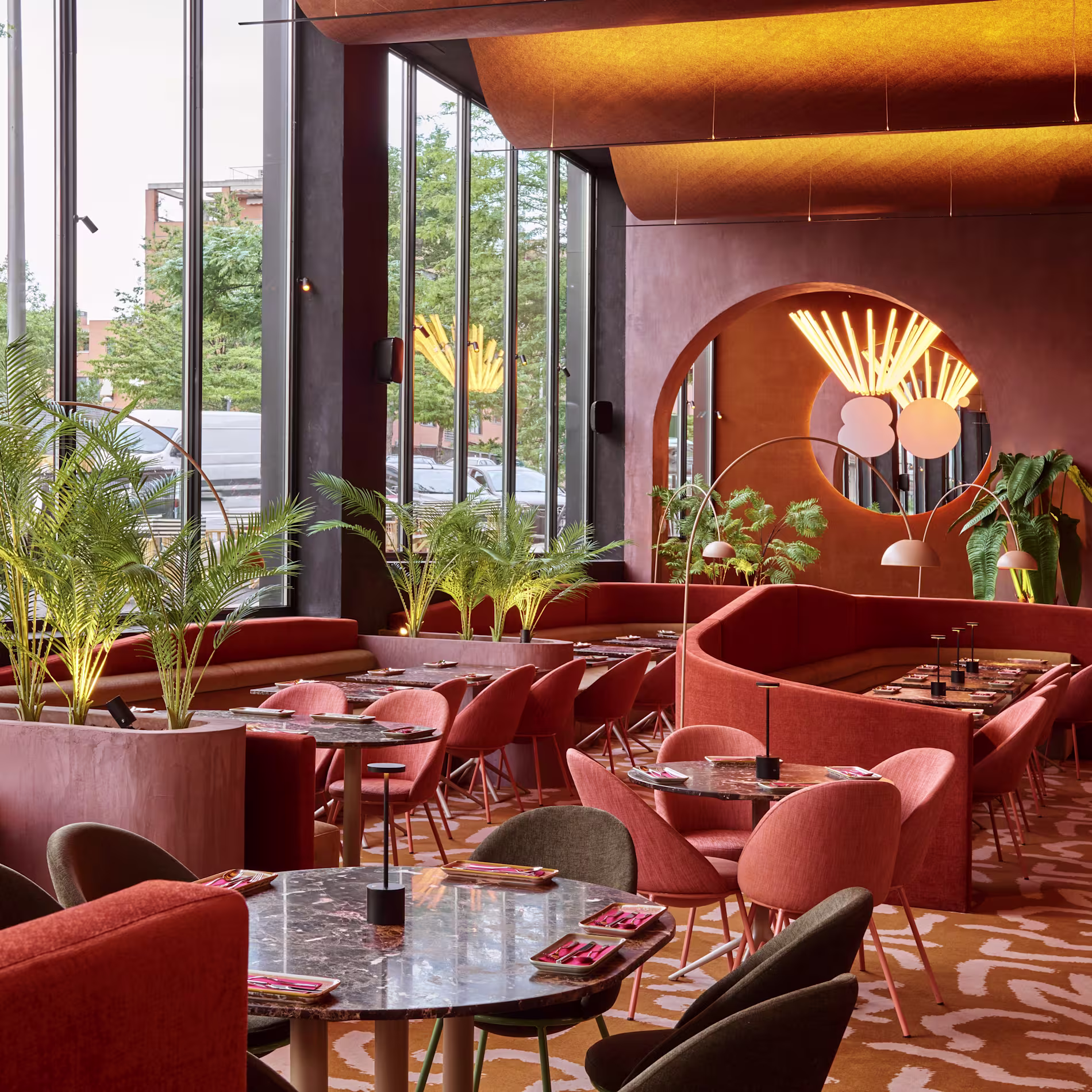 Interior de Ochenta Grados Montecarmelo con grandes ventanales, plantas, tonos cálidos y un diseño moderno con mesas redondas y sofás envolventes.