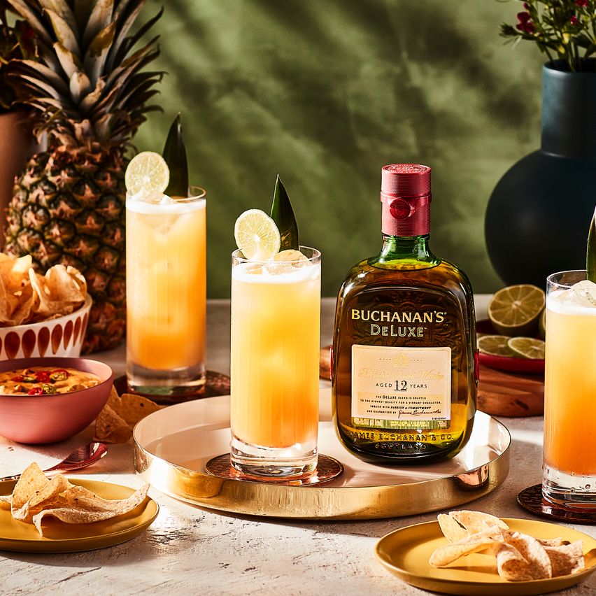 La imagen muestra tres vasos largos con el cóctel Buchanita, una botella de whisky Buchanan's 12 YO y varios platos con alimentos variados.