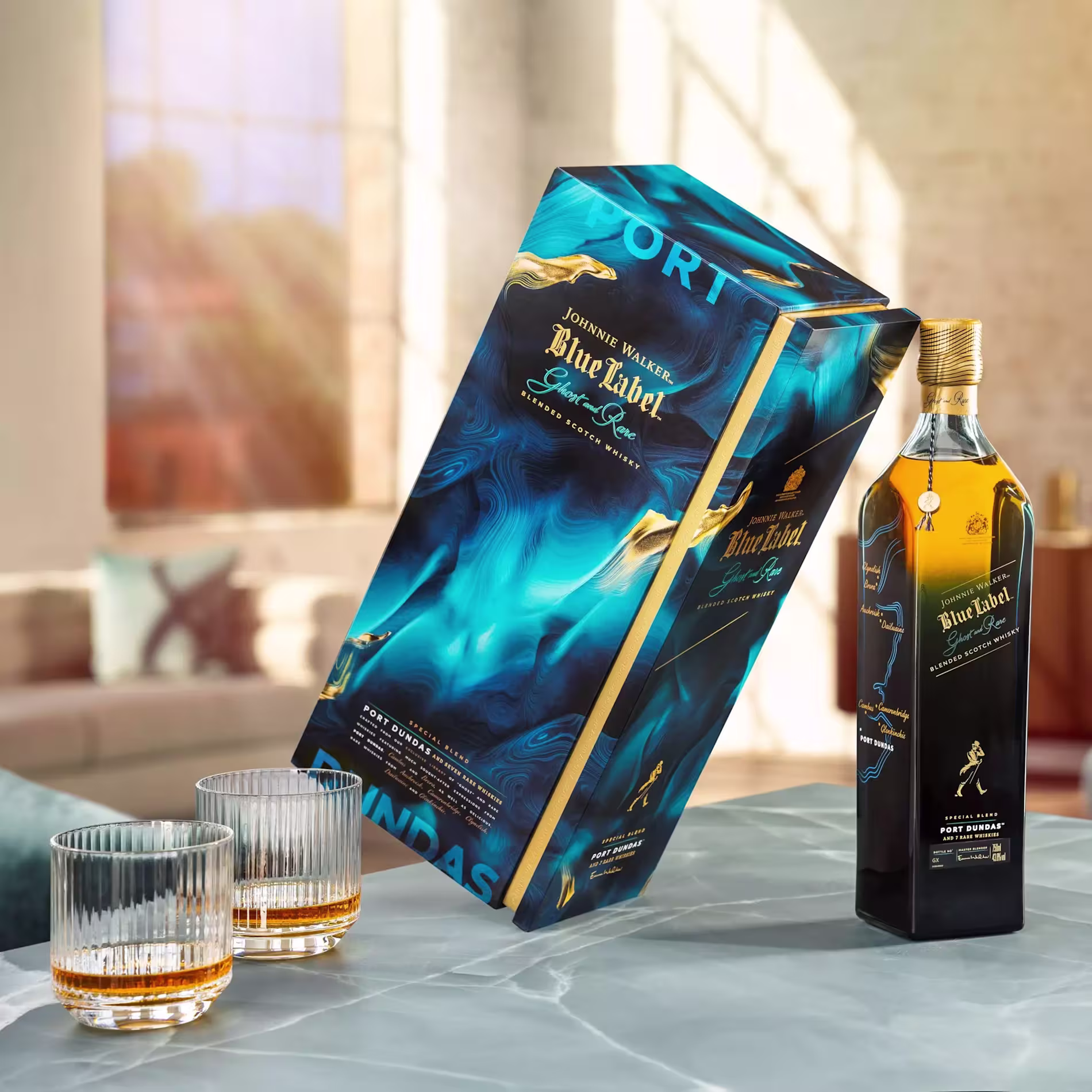 A bottle of Johnnie Walker Blue Label Port Dundas Scotch whisky stands next to its blue and gold box and two whisky glasses on a marble table in a bright, modern room.