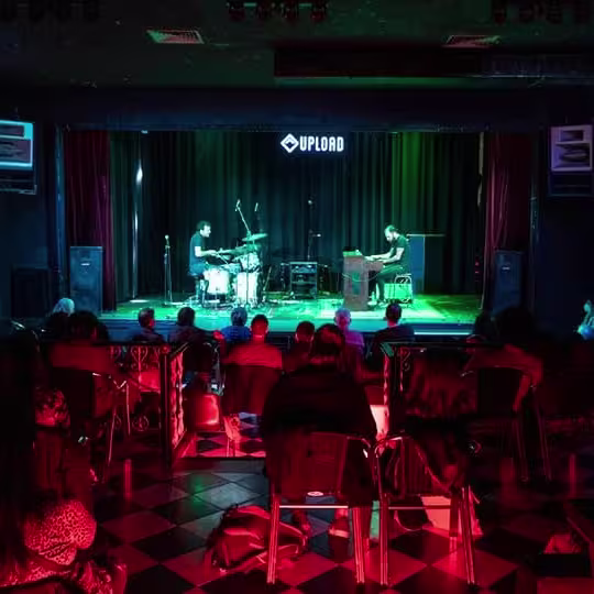Público disfrutando de un concierto íntimo en Sala Upload, con músicos en el escenario y luces verdes y rojas iluminando el espacio.