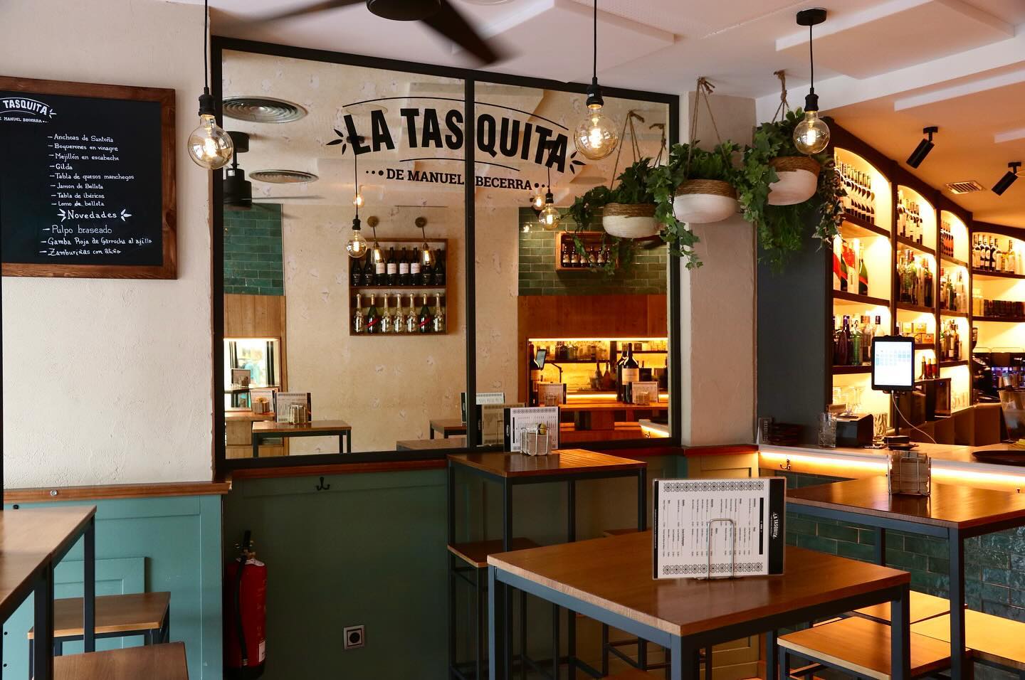 Interior acogedor de La Tasquita, con mesas de madera, estanterías de botellas iluminadas y detalles verdes que crean un ambiente cálido y tradicional.