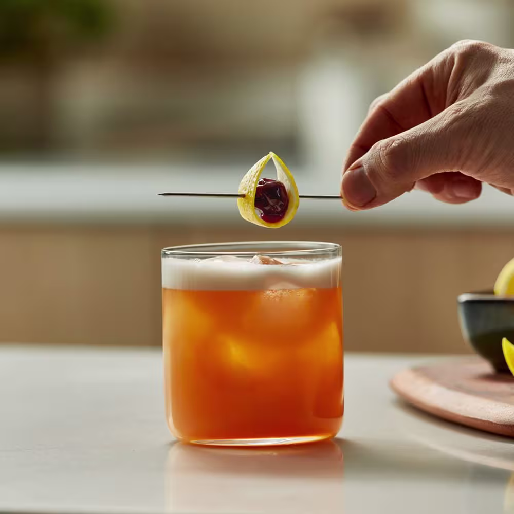 A hand holds a cocktail garnish of a cherry inside a lemon peel above a glass of orange-colored cocktail with ice on a countertop.