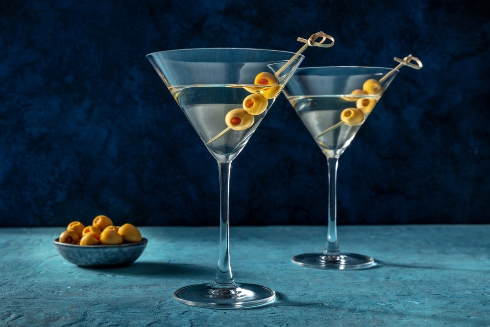 Two martini glasses with olives on cocktail picks, placed on a blue surface with a small bowl of olives in the background.