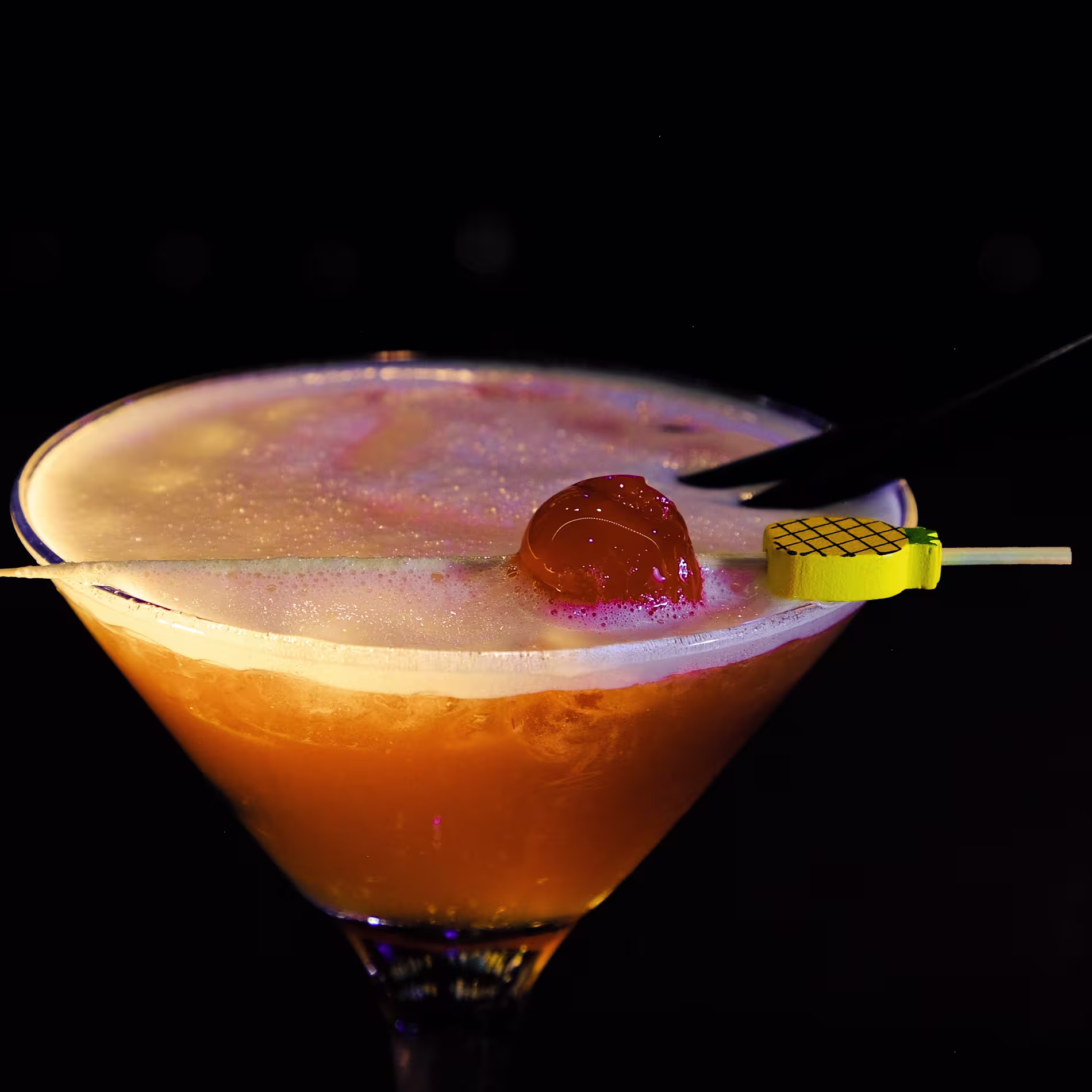 La imagen muestra un primer plano de un cóctel en una copa de martini. La bebida es de color ámbar con una capa espumosa en la parte superior.