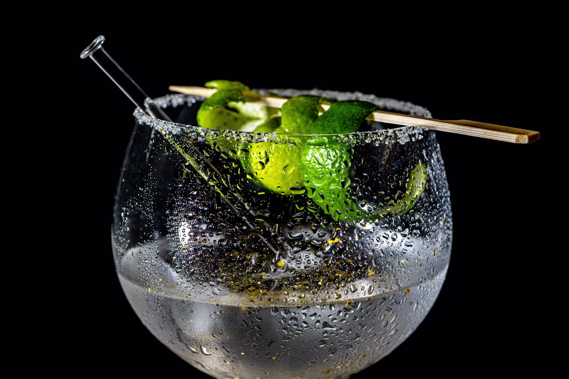 A close-up of a chilled cocktail in a round glass with a salted rim, lime peels, bamboo skewer, and a stirrer, set against a black background.