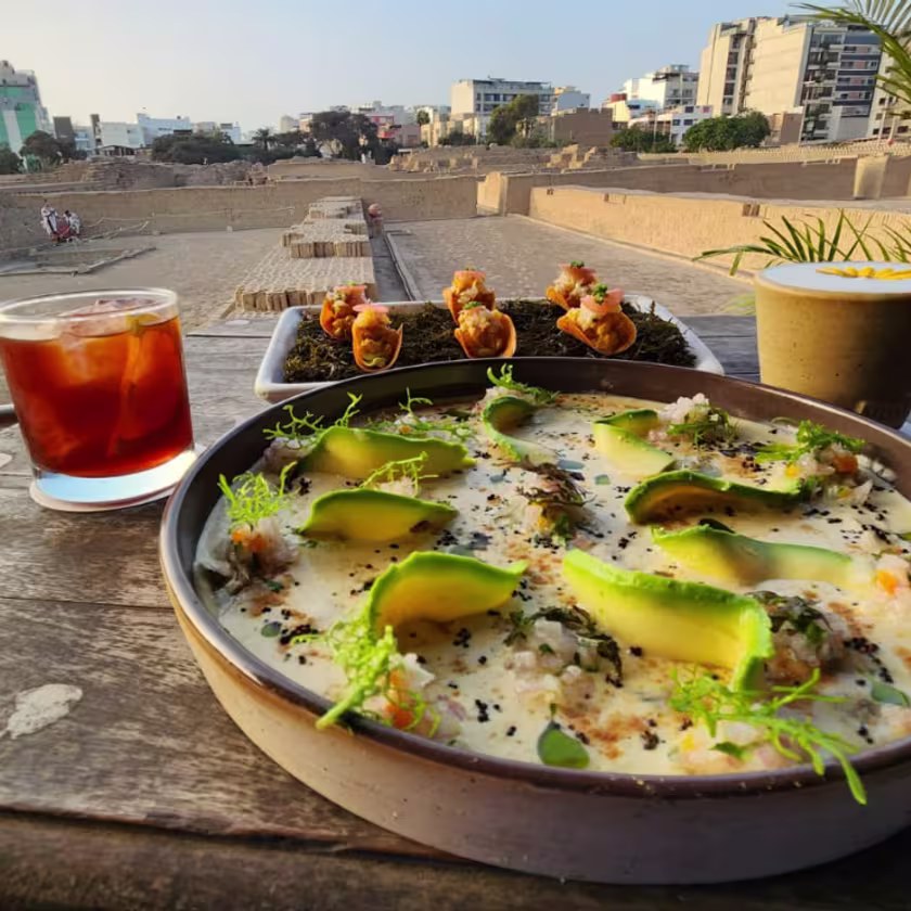 En la imagen se ve un plato de comida, con dos cócteles por detrás, donde también se ve de fondo un paisaje de playa.