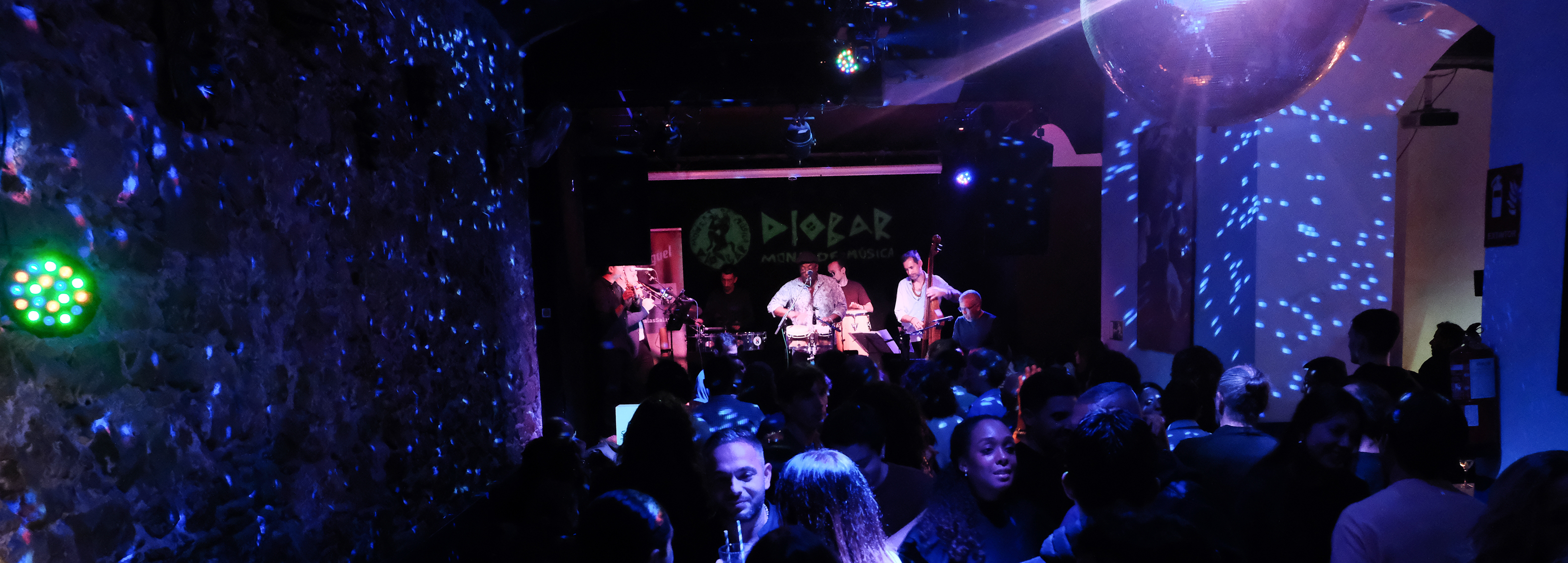La imagen muestra el interior del DIobar  lleno de gente con una banda en vivo actuando en el escenario. La iluminación es tenue con luces de colores y luces de discoteca dispersas por la sala, creando una atmósfera animada. 
