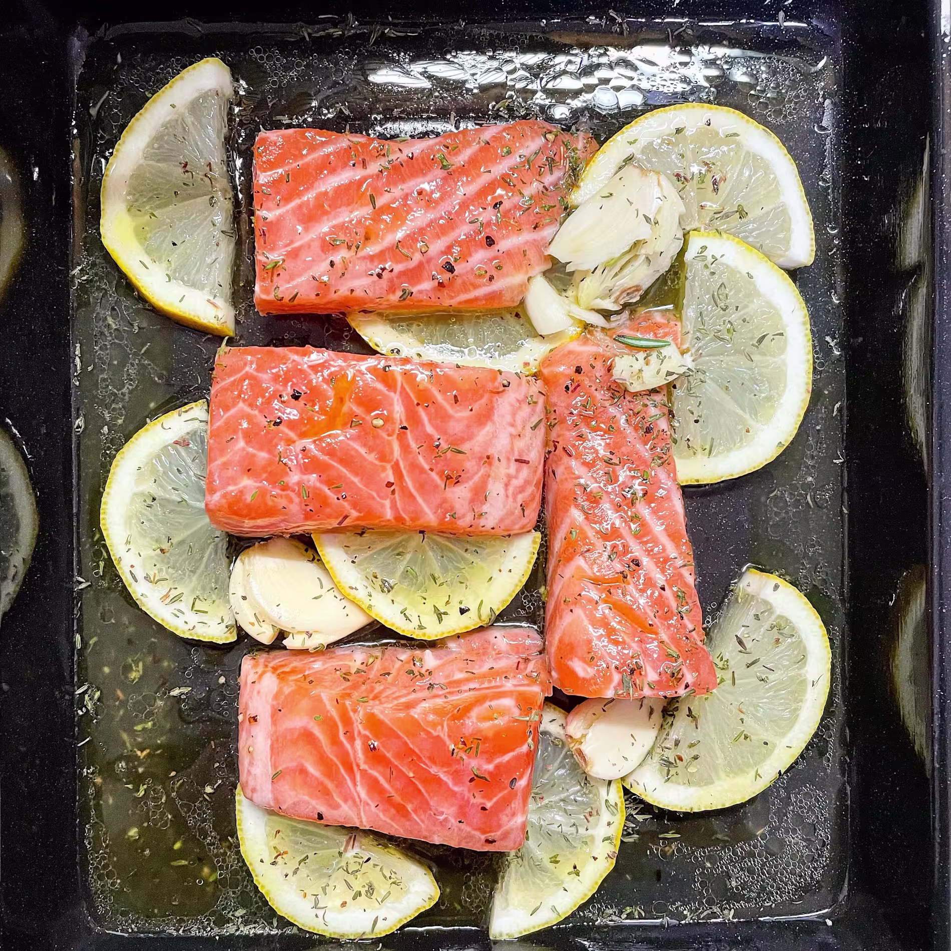 Raw salmon fillets seasoned with herbs are arranged on lemon slices and garlic cloves in a black baking dish.