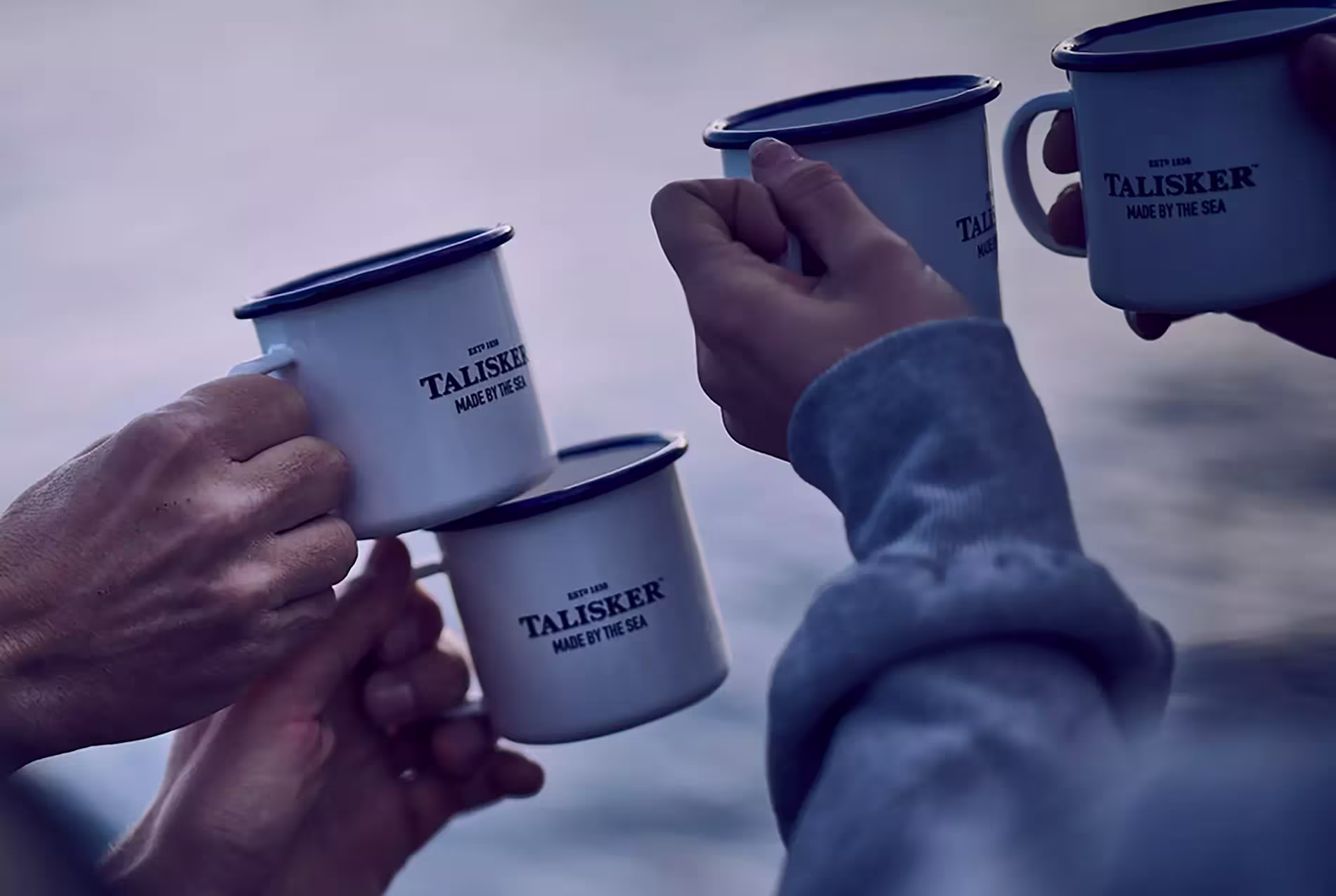 Four people clinking white metal mugs labeled Talisker Made by the Sea against a blurred outdoor background.