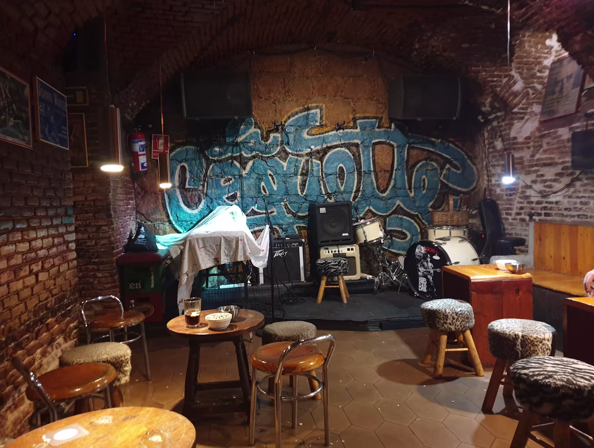 La imagen muestra un espacio interior de La Coquette, con un ambiente rústico y vintage. Las paredes son de ladrillo visto y hay varias mesas y taburetes de madera dispuestos en el suelo. El techo tiene una estructura de ladrillo arqueado.