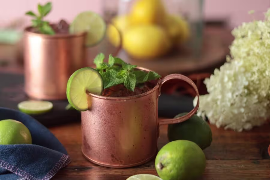 A copper mug filled with a cocktail, garnished with lime and mint, sits on a wooden table with limes, lemons, and flowers in the background.