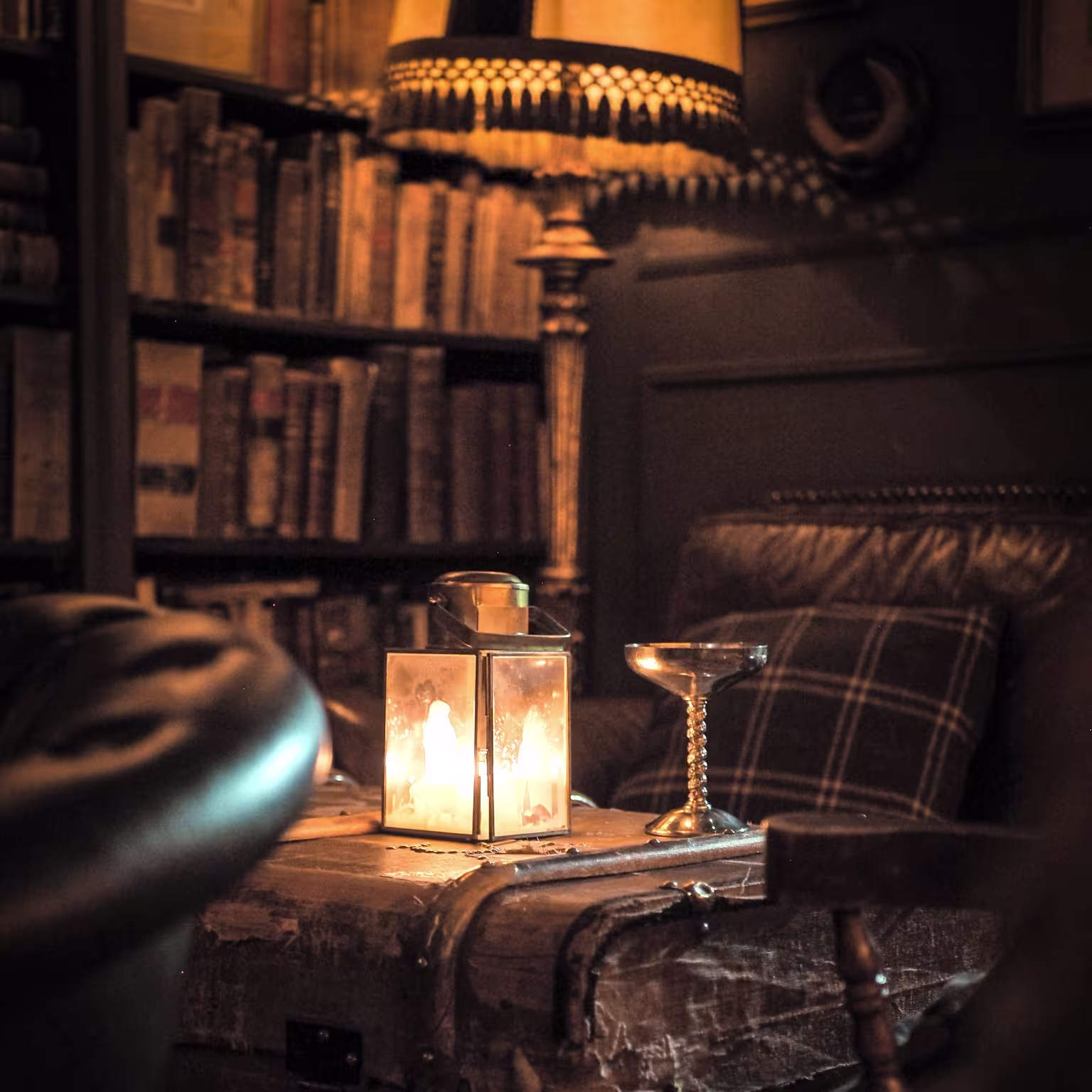 La foto muestra el Jack´s library. En el centro, hay una linterna encendida colocada sobre lo que parece ser un baúl de cuero antiguo. Al lado de la linterna, hay una copa de vino, que refleja la luz. En el fondo, se puede ver un sillón de cuero.