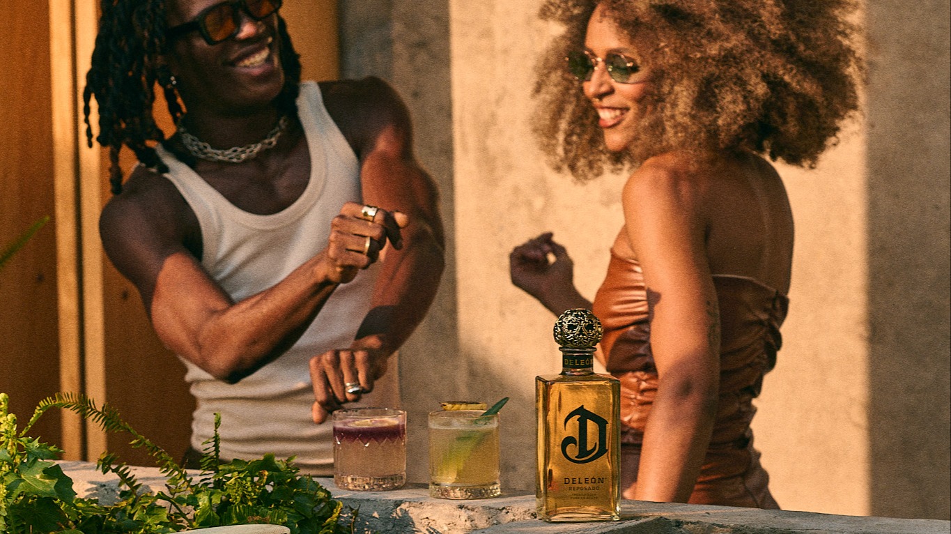 Two people are smiling and dancing near a table with two cocktails and a bottle of DeLeón Tequila outdoors.