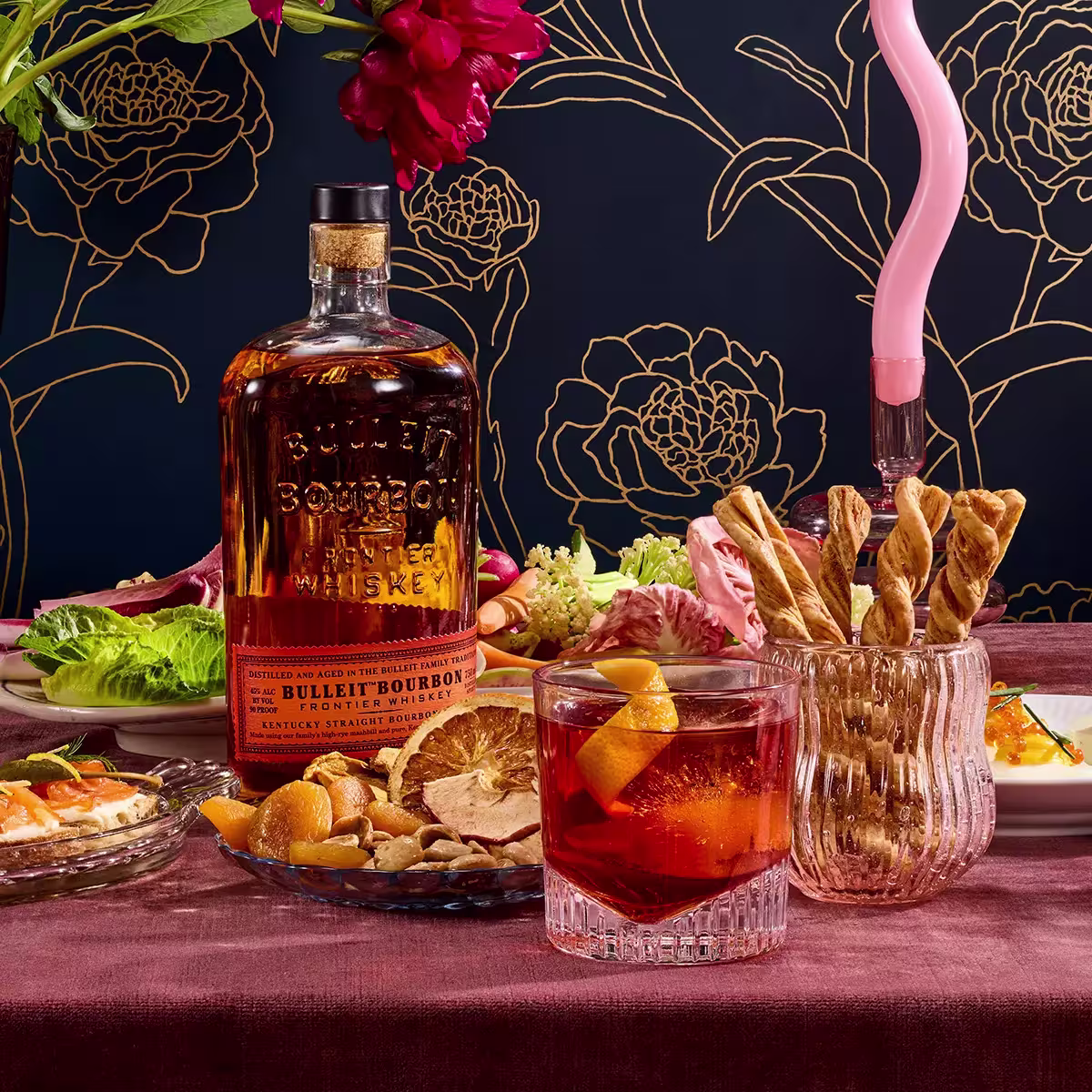 A bottle of Bulleit Bourbon whiskey next to a cocktail, breadsticks in a glass, dried fruit, salad, and other dishes on a table with a dark floral-patterned background.