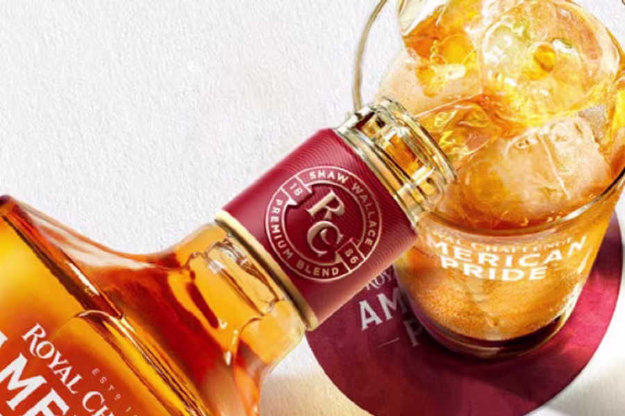 A bottle of Royal Challenge whisky is shown next to a glass filled with whisky, ice cubes, and a coaster labeled American Pride.