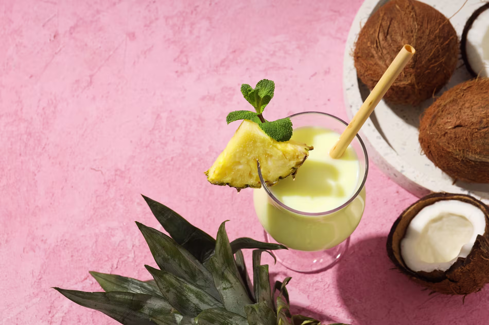 A glass of yellow smoothie garnished with pineapple and mint is placed on a pink surface, next to whole and halved coconuts and a pineapple top.