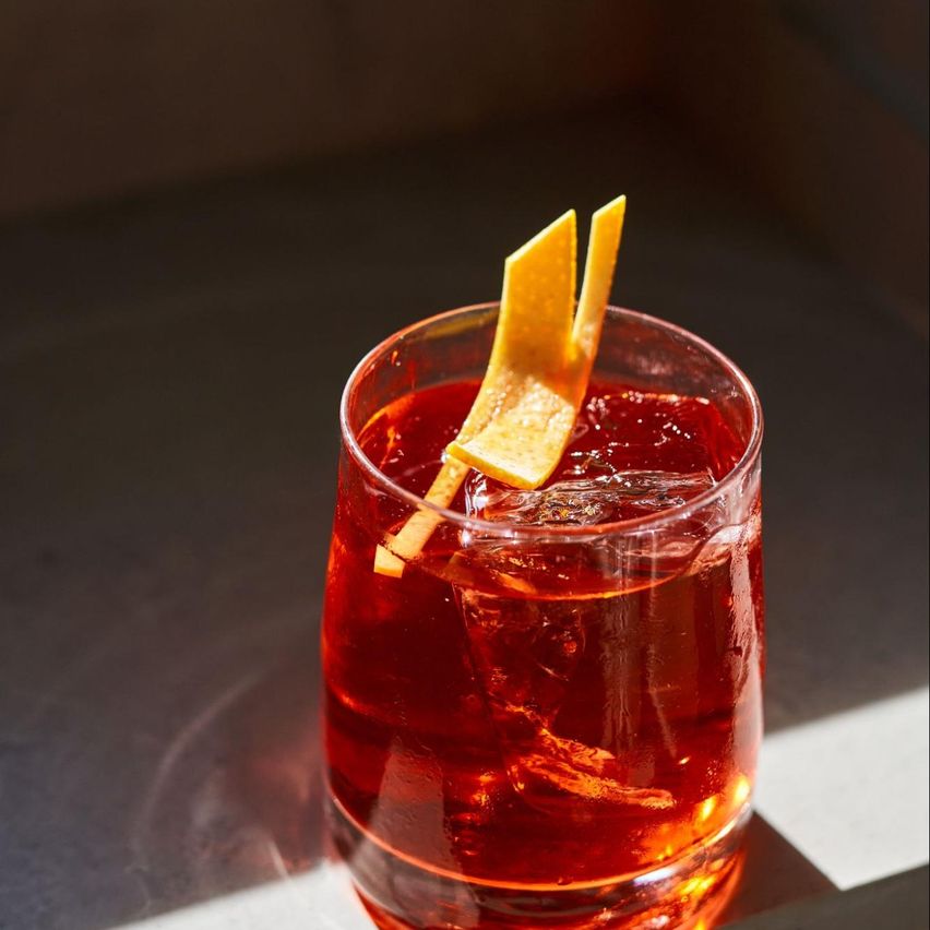 Imagen del trago Negroni, en tono rojo, decorado con rodaja de naranja en al parte superior. La foto esta tomada con un efecto de luz del sol que aporta calidez.