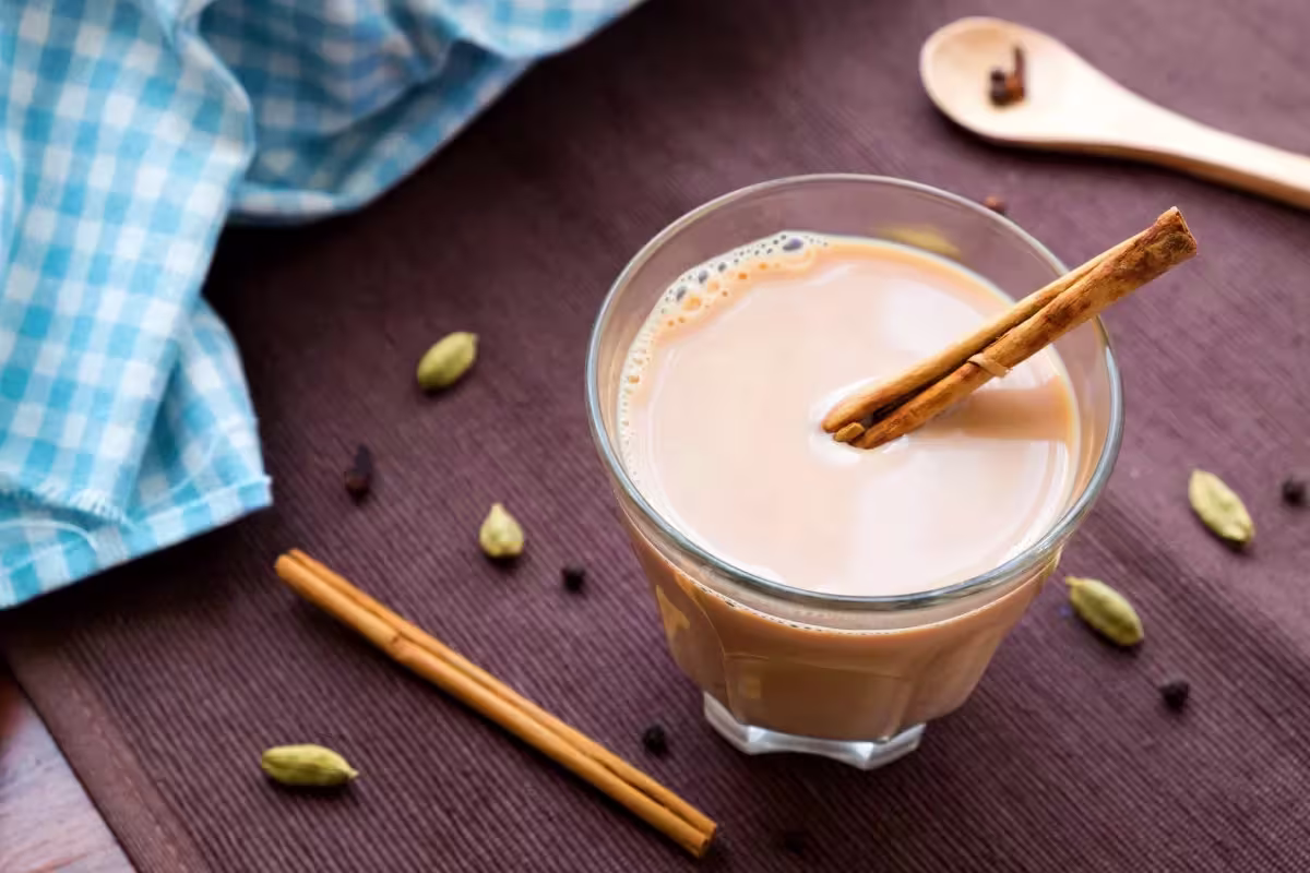 A glass of chai tea with a cinnamon stick, placed on a brown mat with scattered cloves, cardamom pods, a wooden spoon, and a blue-checkered cloth nearby.