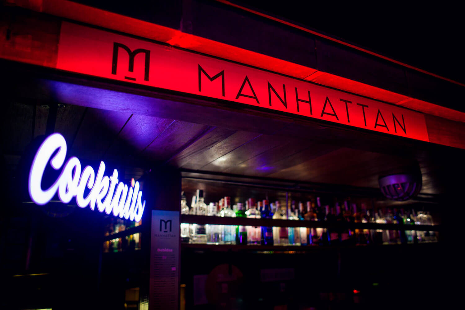 Barra de Manhattan River Bar con letrero rojo iluminado, cartel neón de “Cocktails” y estanterías llenas de botellas de licor.