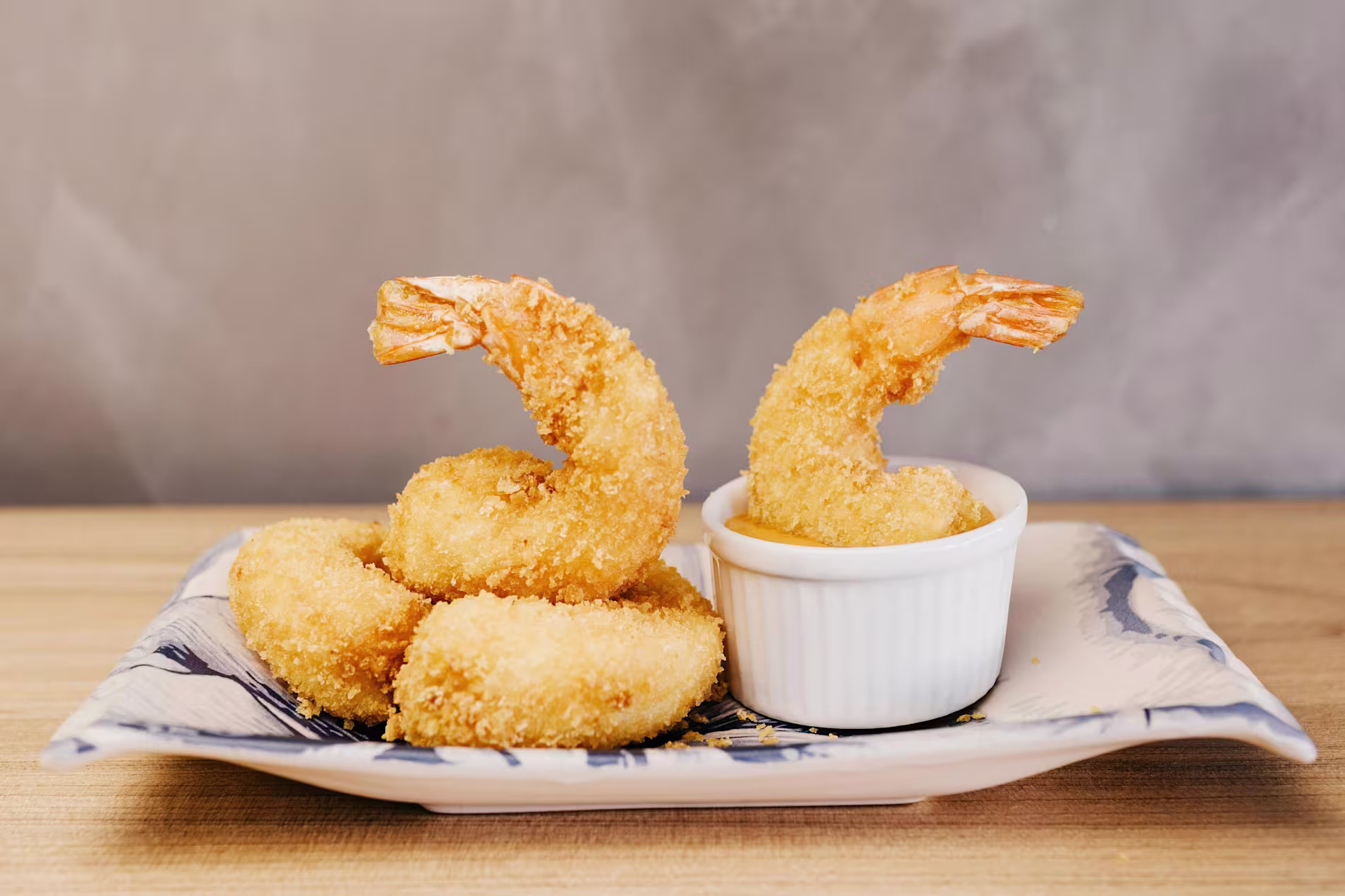 Four pieces of breaded fried shrimp are served on a rectangular plate with a small white ramekin of dipping sauce.