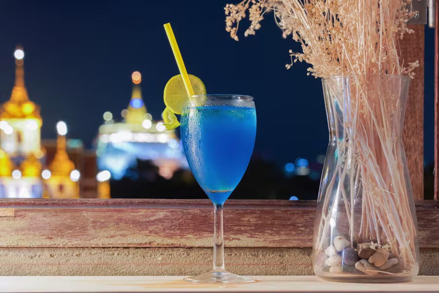 A blue cocktail with a lemon slice and yellow straw sits by a window with city lights in the background and a vase of dried plants beside it.