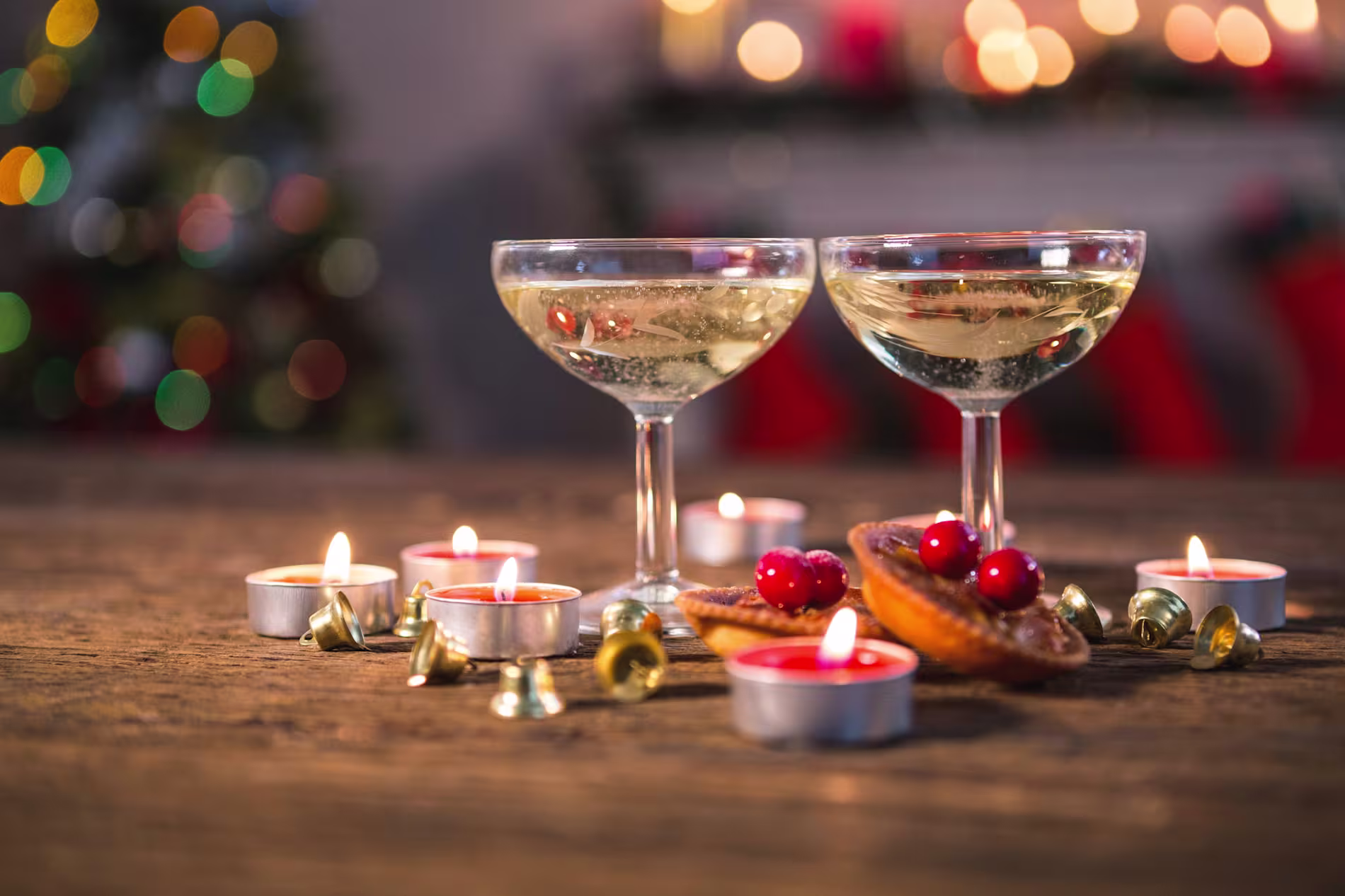 Two glasses of champagne on a wooden table surrounded by lit tealight candles, small bells, mince pies, and red berries, with festive lights blurred in the background.