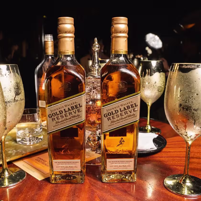 Dos botellas de Johnnie Walker Gold Label Reserve rodeadas de copas doradas en referencia al cóctel Golden Champ.