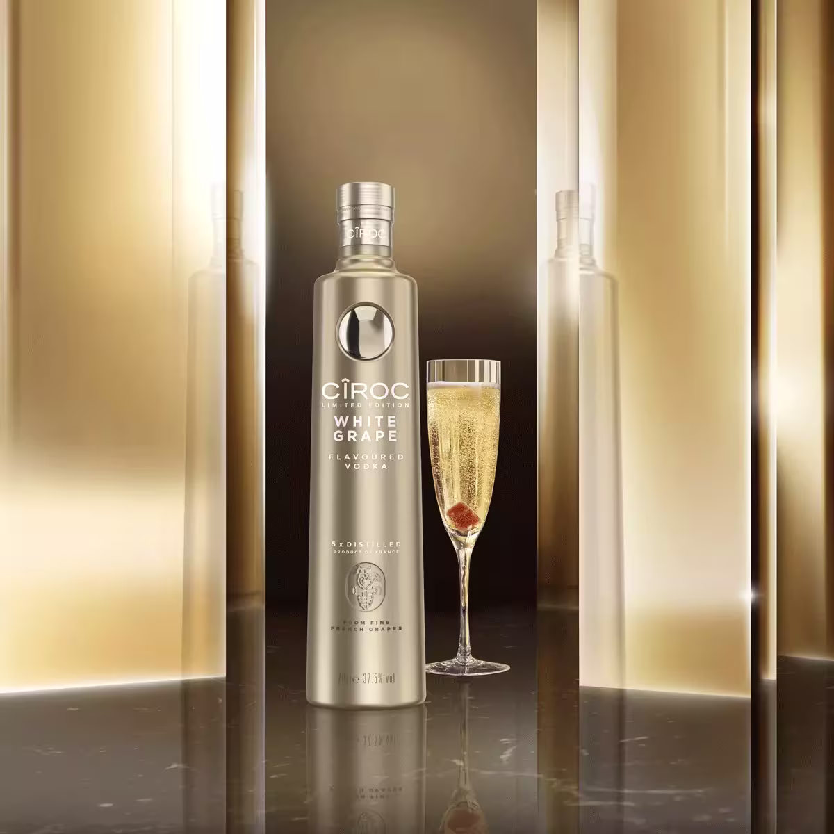 A bottle of Cîroc White Grape vodka stands next to a filled champagne flute on a reflective surface with a gold-toned background.