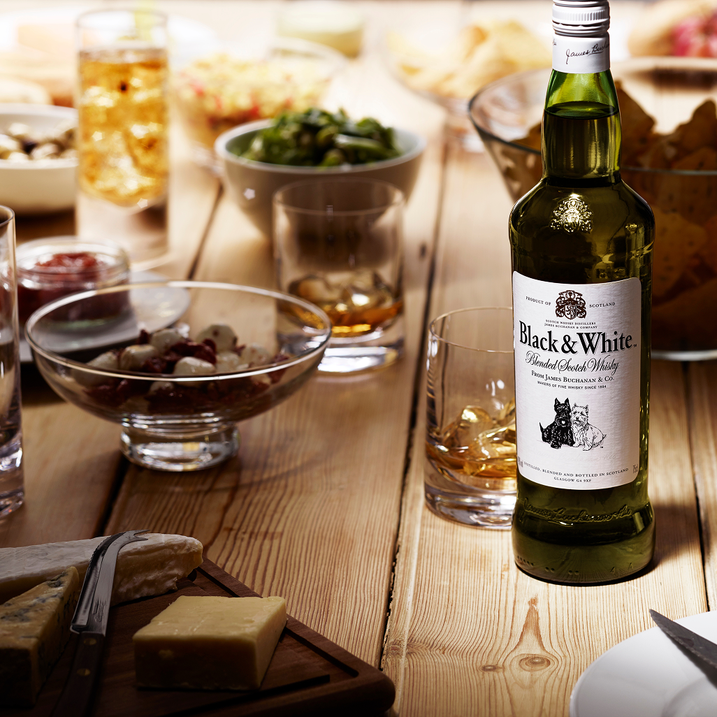 Black & White whisky bottle on a wooden table surrounded by snacks, glasses, and appetizers