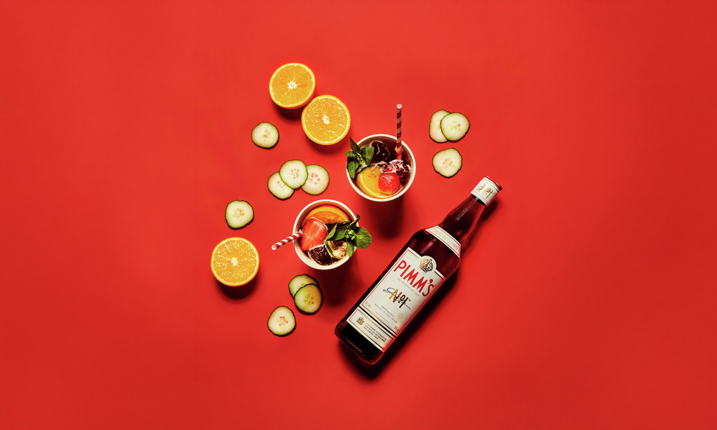 Pimms bottle with a fruit-filled cup, cucumber slices, and an orange half on a vibrant red background