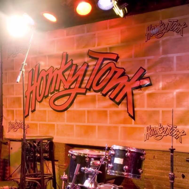 La foto muestra un espacio interior del Honky Tonk. Hay grandes lámparas colgantes blancas que iluminan el área. Las paredes están pintadas en tonos cálidos de naranja y rojo, con cuadros enmarcados colgados en ellas.