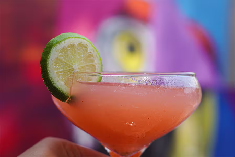 A pink cocktail garnished with a slice of lime on the rim of a coupe glass, with a colorful blurred background.