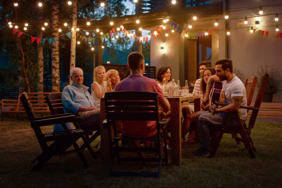 A group of people sit around a table outdoors in the evening under string lights, with one person playing guitar and others listening.