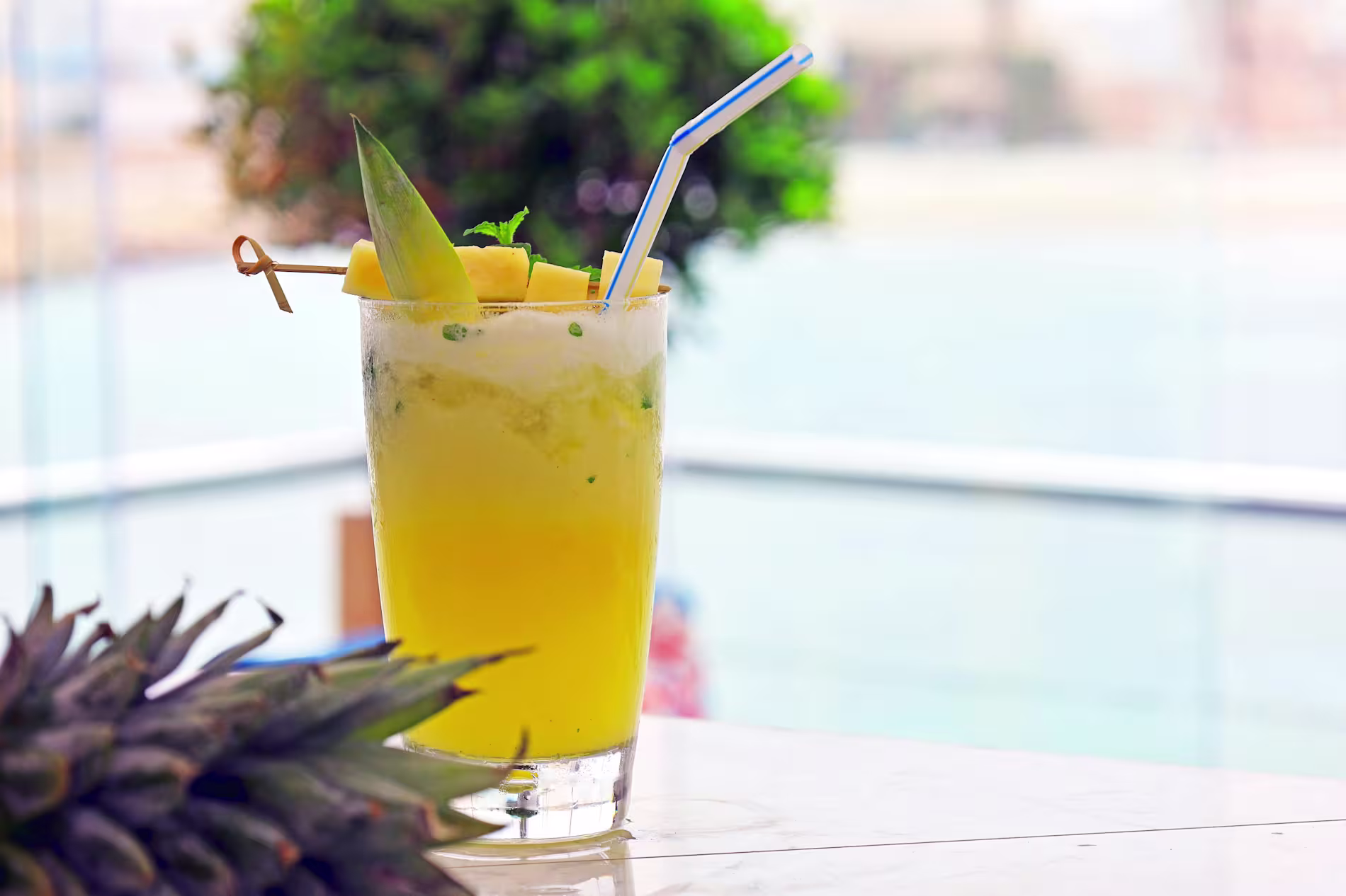 A glass of yellow pineapple juice with ice, mint, pineapple garnish, and straw sits on a white table with blurred outdoor background.