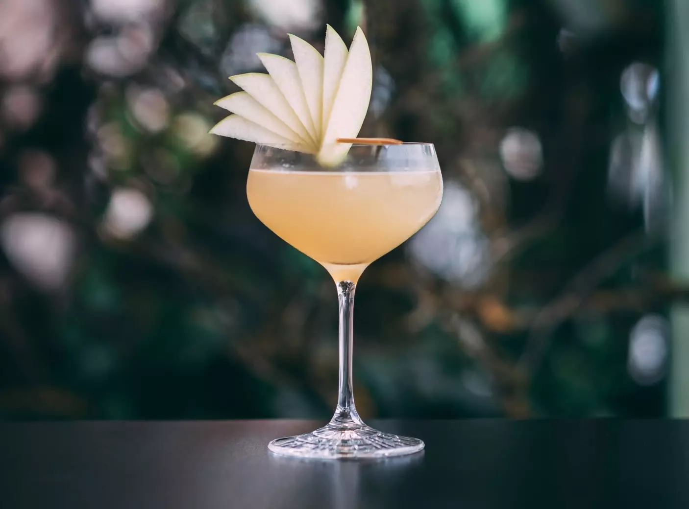 A stemmed cocktail glass filled with a light yellow drink, garnished with thinly sliced apple fan on the rim, set against a blurred background.