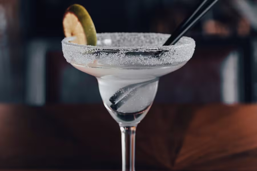 A margarita glass with a salted rim, filled with a clear beverage, garnished with a lime wedge, and two black straws.