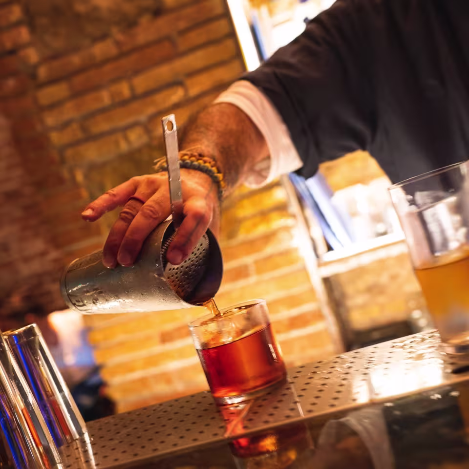 Un bartender sirve un cóctel ámbar con coctelera y colador sobre la barra, en un ambiente cálido de ladrillo visto y luz tenue.