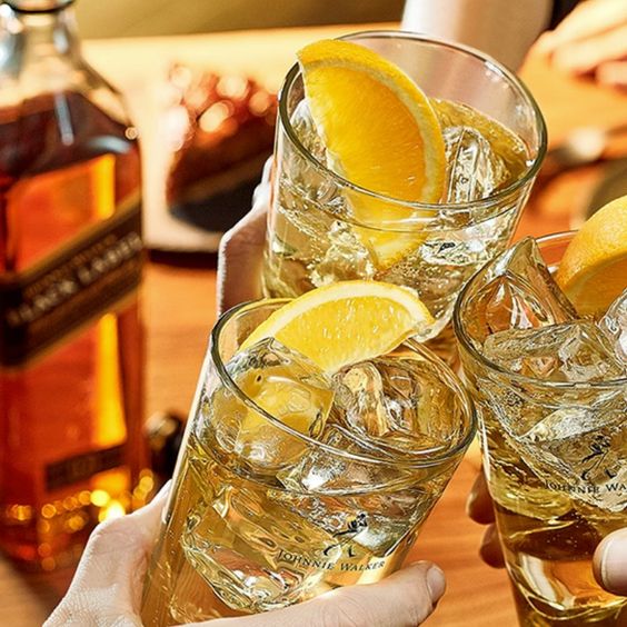 La imagen muestra un brindis con tres vasos con cócteles preparados con whisky Johnnie Walker y de fondo, una botella de whisky Johnnie Walker Black Label y distintos platos de comida.