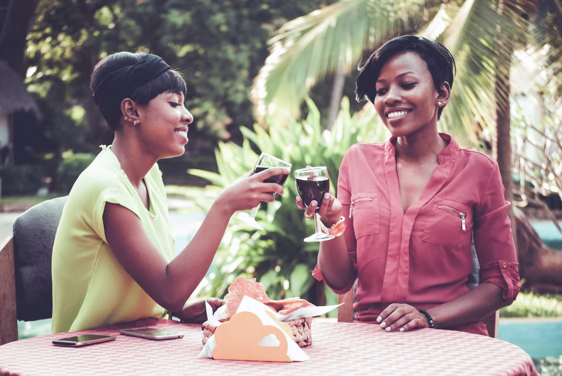 Two women sit at an outdoor table, smiling and clinking glasses of red wine. Food and smartphones are on the table, and greenery is visible in the background.