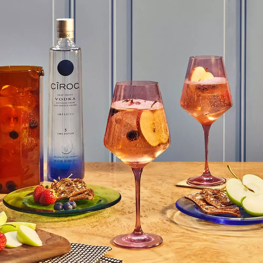 A bottle of Cîroc vodka, two pink stemmed glasses with fruit cocktails, plates of dried fruit, fresh apple slices, and berries on a wooden table.