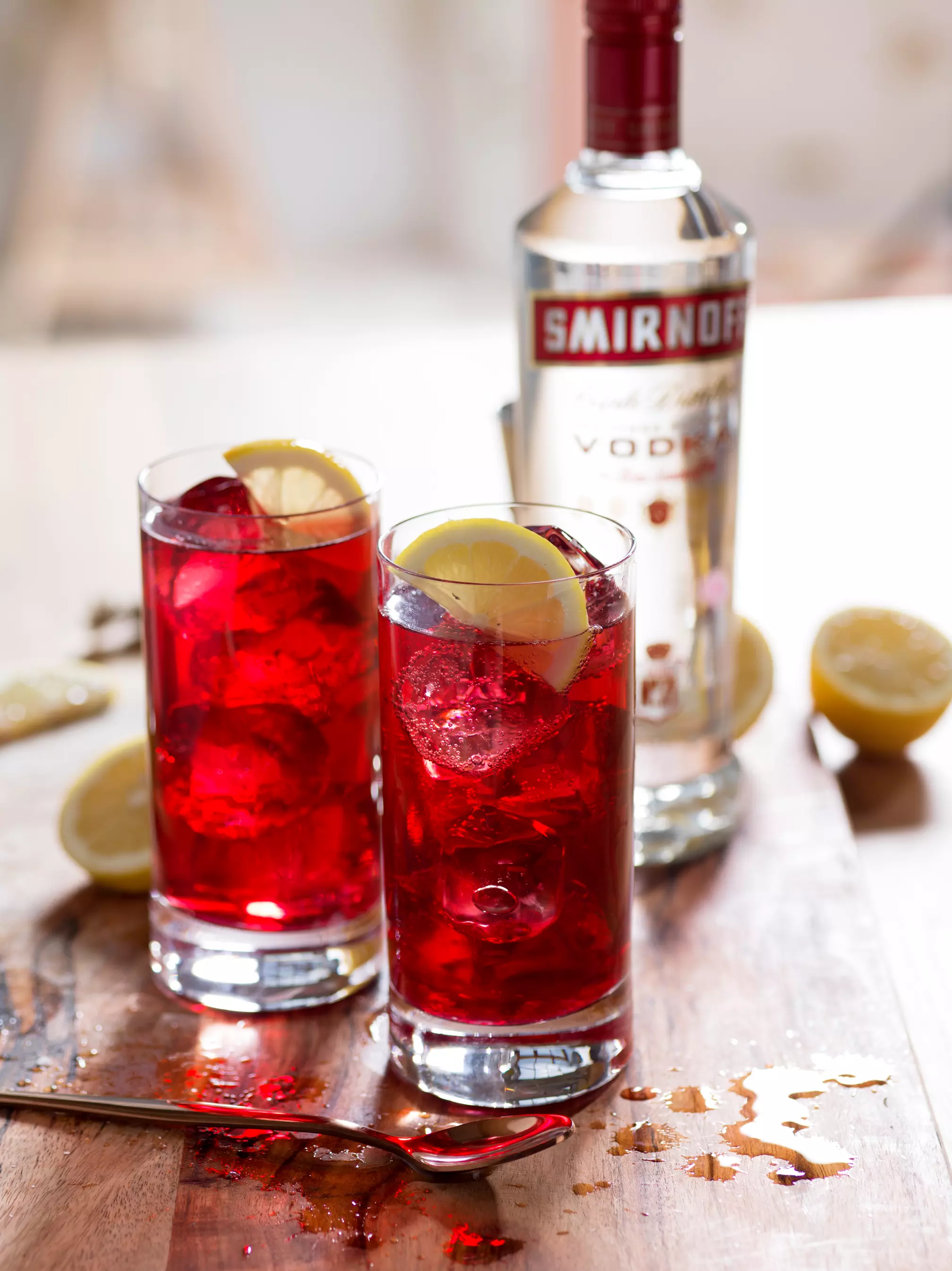 Two tall glasses filled with red cocktails, ice, and lemon slices are on a wooden surface next to a bottle of Smirnoff vodka and a sliced lemon.