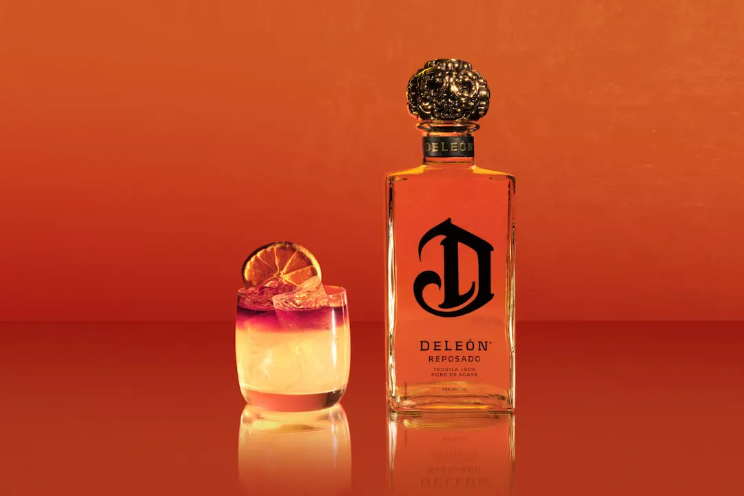 A bottle of DeLeón Reposado tequila stands next to a cocktail garnished with a dried citrus slice against an orange background.
