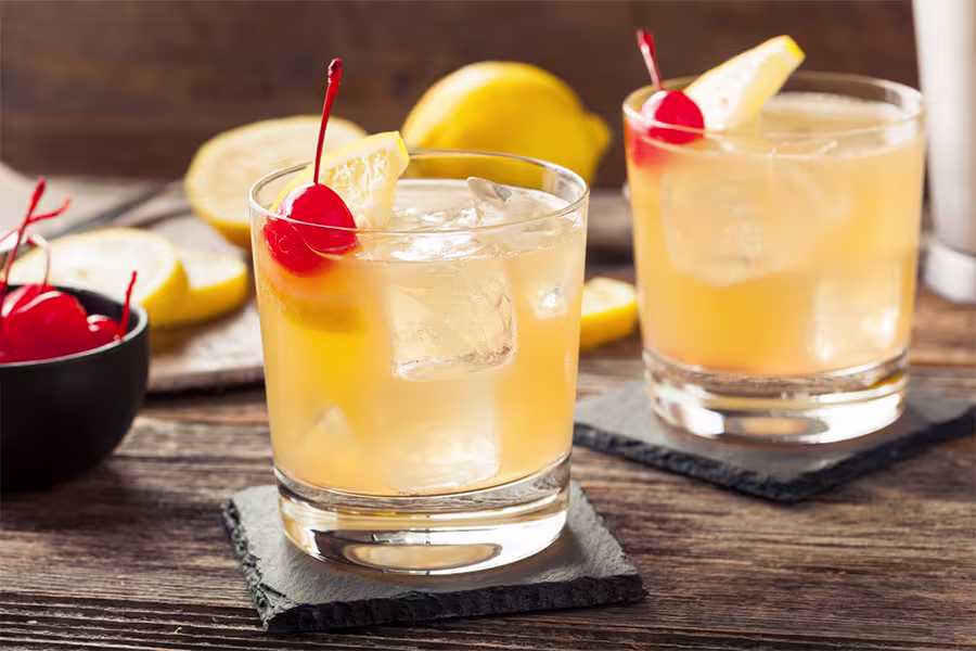 Two whiskey sour cocktails with ice, garnished with lemon slices and maraschino cherries, on slate coasters with lemons and cherries in the background.