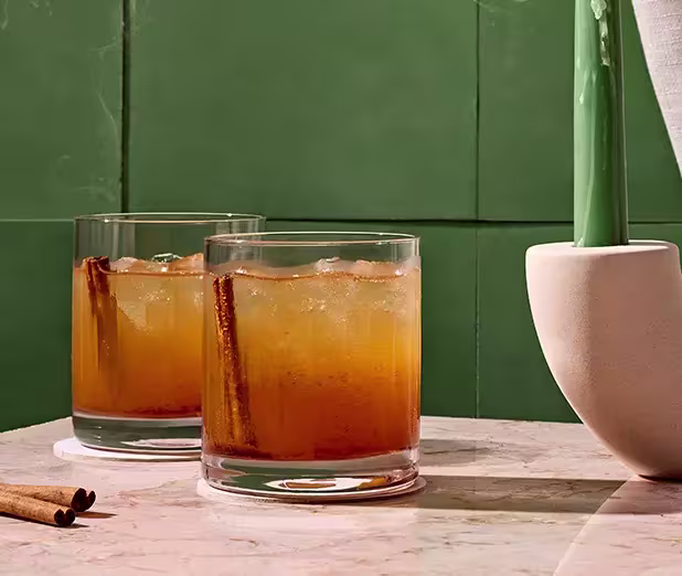 Two short glasses with an amber-colored drink and ice, each garnished with a cinnamon stick, placed on a marble surface next to a vase, with a green tiled background.